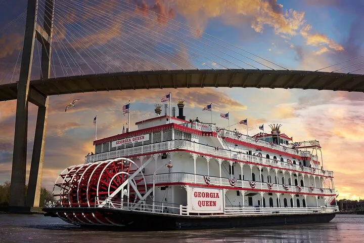 Georgia Queen riverboat sailing under a large suspension bridge at sunset.